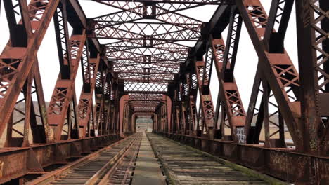 Camera-panning-down-over-an-old-rail-road-bridge-over-a-river-in-an-industrial-town