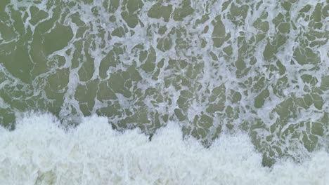 Drone-descends-to-foamy-whitewash-from-behind-wave-crashing-on-empty-beach,-aerial