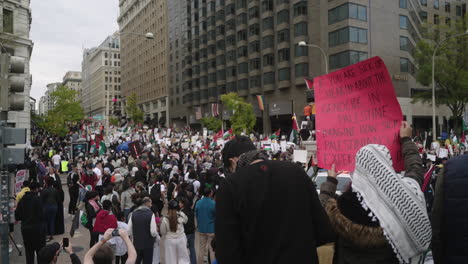 A-Protestor-in-a-Keffiyeh-Holds-up-a-Pro-Palestine-Sign-with-a-Large-Crowd-of-Protestors-Marching-in-the-Streets-in-Front-of-Them,-wide-shot