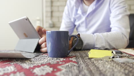 Hombre-De-Negocios-Tocando-La-Tableta-Con-Taza-De-Café-Y-Llaves-En-La-Mesa,-Trabajando-Desde-Casa