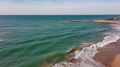 Port-Ginesta-Con-Aguas-Azules-Y-Yates-Amarrados,-Suaves-Olas-Rompiendo-En-La-Playa-De-Arena,-Día-Soleado,-Vista-Aérea