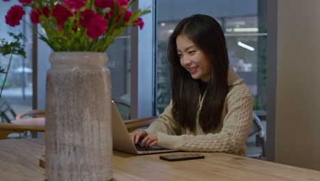 Happy-young-Asian-girl-in-good-mood-working-online-on-her-laptop-from-cafe,-smiling