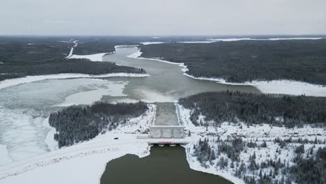 Drones-De-Gran-Altitud-En-Una-Larga-Carretera-Cubierta-De-Nieve-Con-Agua-Corriendo-Por-La-Presa-Hidroeléctrica-Notigi-En-El-ártico