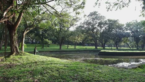 Sun-dappled-park-with-girl-by-the-lake,-green-trees,-tranquil-atmosphere,-daylight