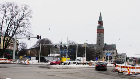 Cityscape-of-Helsinki-with-cars,-tram,-and-pedestrians,-overcast-day,-construction-site-foreground,-with-historic-Helsinki's-National-Museum-building-in-the-background