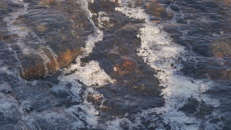 Schmelzwasser-Fließt-über-Die-Dunklen-Felsen,-Die-Mit-Einer-Dünnen-Schicht-Aus-Eisfiligran-Bedeckt-Sind