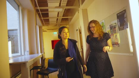 Two-business-woman-walking-through-office-buidling-corridor-entering-meeting-room