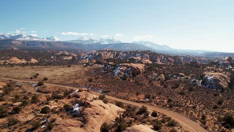 A-high-flying-drone-shot-over-a-remote-dirt-road-cutting-through-the-vast-and-unique-desert-land-near-Moab,-Utah,-with-the-snowy-Rocky-Mountains-towering-in-the-distance