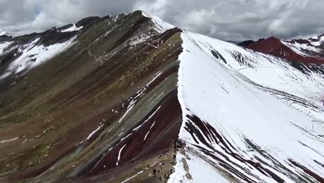 Drone-view-in-Peru-flying-over-rainbow-mountain-in-Cuzco,-showing-a-mountain-with-half-of-it-covered-in-snow-and-the-other-different-red-colors