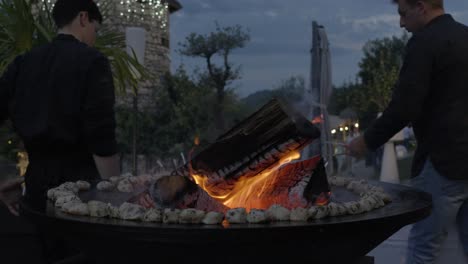 Big-bonfire-with-firewood-and-red-flame-at-wedding-party,-marshmallows-roasting-around