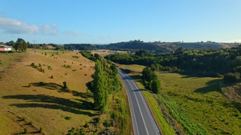 Bird's-eye-view-of-a-passing-car-on-a-rural-road-with-meadows-at-sunset,-Chiloe,-Chile