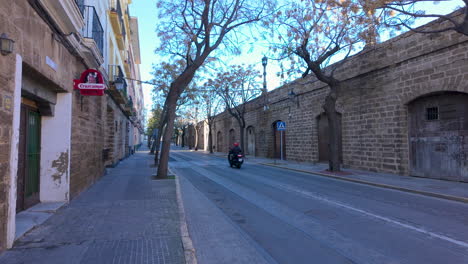 Una-Vista-Desde-Un-Tuktuk-Muestra-Una-Calle-Tranquila-En-Cádiz,-Con-Un-Motociclista-Pasando-Por-Edificios-Tradicionales-De-Piedra-Y-Un-Cielo-Azul-Claro-Arriba.