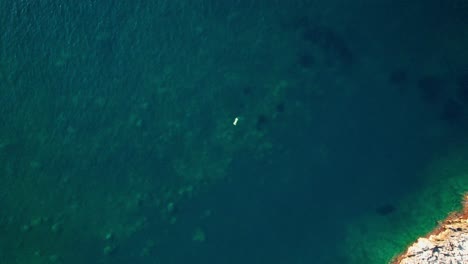 Bird's-eye-view-of-boat-floating-in-middle-of-deep-blue-ocean-water-with-rocky-surface-below