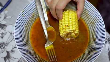 Corn-Cob-Being-Dipped-In-Spice-Sauce-Inside-Bowl