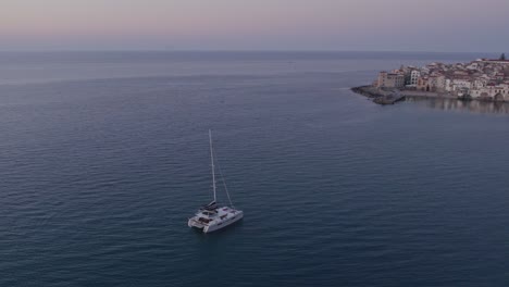Vista-Aérea-Del-Barco-En-El-Mar-En-Calma-Con-Gente-Durante-La-Puesta-De-Sol,-Cefalu,-Sicilia,-Italia