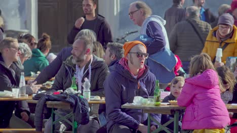 People-sit-on-benches-at-a-table-during-a-festival-and-consume-food-and-drink-while-talking-to-each-other-and-other-people-walking-past-them