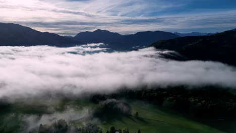 Una-Impresionante-Vista-Aérea-De-Un-Valle-Brumoso,-Con-La-Luz-Del-Sol-Atravesando-Las-Nubes-E-Iluminando-Los-Picos-De-Las-Majestuosas-Montañas-Al-Fondo