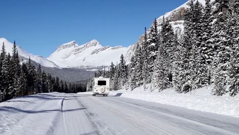 Camper-van-driving-down-a-snowy,-icy-winter-road-surrounded-by-snow-covered-mountains