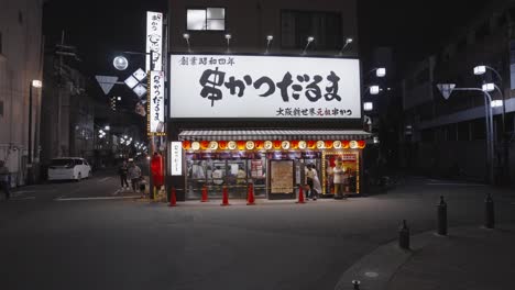 Slow-moving-shot-through-dark-streets-at-night-towards-brightly-lit-Kushikatsu-Daruma-restaurant-Ebisuhigashi,-Naniwa-Ward,-Osaka,-Japan