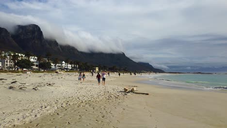 Slo-mo-clip-of-people-and-dogs-walking-on-Camp's-Bay-Beach,-as-clouds-cover-hills-in-the-background