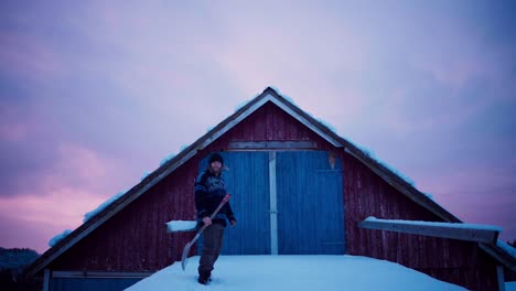 Hombre-Con-Pala-Caminando-Sobre-El-Techo-Cubierto-De-Nieve-Hacia-La-Entrada-Al-ático-De-La-Cabaña-De-Madera-Al-Atardecer