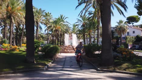 Young-man-in-sunglasses-rides-bike-through-park-with-fountain,-France