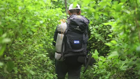 Plano-Medio-Sigue-La-Cámara-De-Un-Hombre-Latino-Abriéndose-Camino-A-Través-De-La-Densa-Jungla-Tropical-Con-Un-Machete-Mientras-Usa-Una-Gran-Mochila-De-Senderismo-Con-Un-Parche-De-La-Bandera-Nicaragüense-En-Cámara-Lenta