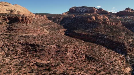 A-high-flying-drone-shot-over-a-remote-dirt-road-cutting-through-the-vast-and-unique-desert-land-near-Moab,-Utah,-with-the-snowy-rocky-mountains-towering-in-the-distance