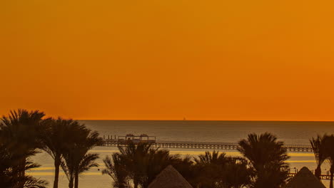 Orange-sunset-over-a-tranquil-sea-with-silhouettes-of-palm-trees-and-a-pier,-timelapse