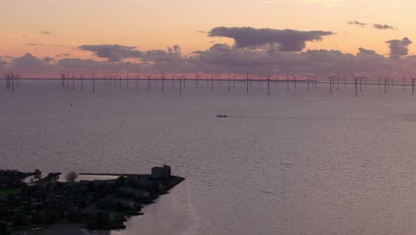 Aerial-view-of-sailing-vessel-with-In-the-background-big-windfarm-with-turbines-at-sunset,-IJsselmeer,-Netherlands