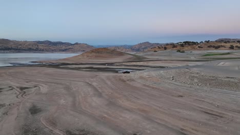 Camioneta-Negra-Conduciendo-A-Lo-Largo-Del-Lecho-De-Un-Lago-Seco-Al-Atardecer-Con-Las-Luces-Encendidas-En-El-Lago-Millerton-Fresno,-California,-60-Fps