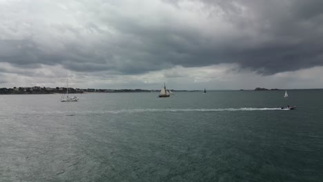 Drone-flying-towards-Le-Renard-Robert-Surcouf-wooden-boat-on-sea-with-cloudy-sky