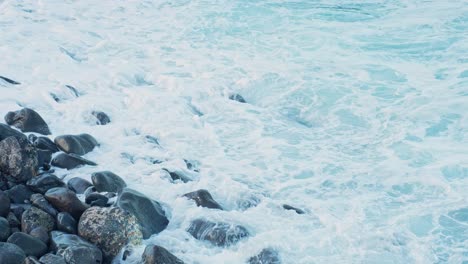 waves-of-water-crashing-towards-the-pebble-stones-on-the-seashore,-static-closeup-slow-motion