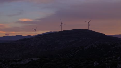 Wind-turbines-spin-on-top-of-exposed-ridgeline-at-blue-hour-with-soft-fire-glow-from-sunset-against-clouds