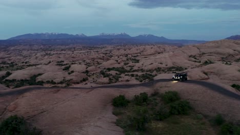 Eine-4K-Drohnenaufnahme-Eines-Hochgefahrenen-Jeeps,-Der-Im-Gelände-Durch-Die-Extreme-Und-Felsige-Wüstenlandschaft-In-Der-Nähe-Von-Moab,-Utah,-Fährt,-Mit-Den-Schneebedeckten-Rocky-Mountains-In-Der-Ferne