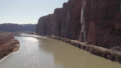 A-tracking-drone-shot-of-a-truck-driving-between-the-steep-cliffs,-known-as-the-“Wall-Street-Climbing-area”,-and-the-Colorado-River,-near-Moab,-Utah