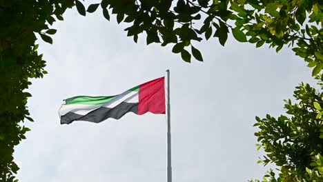 Die-Flagge-Der-Vereinigten-Arabischen-Emirate-Weht-Anmutig-Im-Bogen-Aus-Grünen-Pflanzen-An-Der-Corniche-Von-Abu-Dhabi-Und-Verkörpert-Das-Konzept-Einer-Grünen-Zukunft-In-Den-Vereinigten-Arabischen-Emiraten