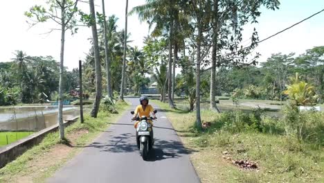 Toma-Aérea-De-Seguimiento-De-Una-Turista-Vestida-De-Amarillo-Montando-Ciclomotor-A-Lo-Largo-De-La-Carretera-En-Canggu,-Bali.
