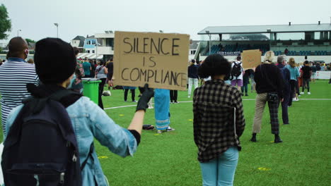 Vista-Posterior-De-Un-Joven-Manifestante-Sosteniendo-Un-Cartel-Que-Dice-Que-El-Silencio-Es-Cumplimiento.