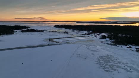 Ein-Schneebedeckter-Hafen-Bei-Sonnenuntergang-Mit-Goldenem-Himmel-Und-Spärlichen-Bäumen,-Luftaufnahme
