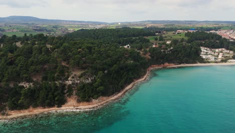 Aerial-shot-of-coastline-with-pine-trees-and-turquoise-water-in-Kallithea,-Halkidiki,-Greece