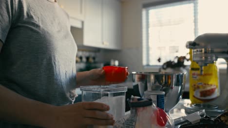 A-woman-measures-and-pours-sugar-into-a-metal-mixing-bowl-in-a-handheld-slow-motion-shot,-featuring-a-slight-push-in-as-she-pours