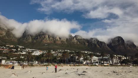 Pan-along-Table-Mountain-and-Camp's-Bay-from-the-beach,-as-clouds-partly-cover-the-hills