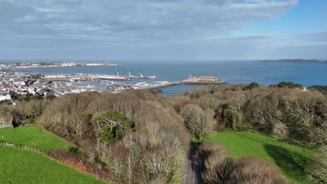 St-Peter-Port-Guernsey-flight-from-top-of-Vals-de-Terres-over-tree-tops-towards-Castle-Cornet-with-views-over-harbour,-marinas,-Belle-Greve-Bay-and-across-to-Herm