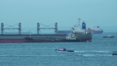 Pequeños-Barcos-De-La-Autoridad-Gubernamental-Navegando-Alrededor-De-Los-Grandes-Buques-De-Carga-En-El-Estrecho-De-Singapur,-Inspeccionando-Los-Productos-Aduaneros-Y-Documentándolos-Antes-De-Que-Lleguen-Al-Interior,-Primer-Plano