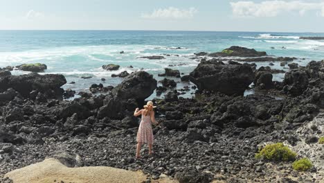 Woman-walking-on-black-rocks-near-the-seashore-while-wearing-floral-dress,-static-slow-motion