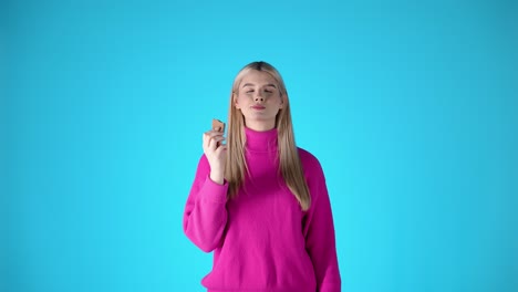 Caucasian-young-blonde-woman-eats-a-cookie-in-a-pink-sweater-chroma-infinite-studio-torso-shot,-blue-background,-smiling-rubbing-her-belly