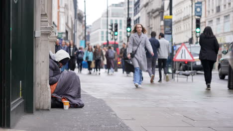 In-slow-motion-people-with-shopping-bags-walk-past-a-homeless-man-sat-against-a-wall,-wrapped-up-in-a-sleeping-bag-on-Oxford-Street