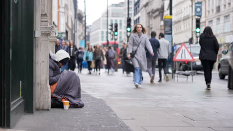 En-Cámara-Lenta,-Personas-Con-Bolsas-De-Compras-Pasan-Junto-A-Un-Hombre-Sin-Hogar-Sentado-Contra-Una-Pared,-Envuelto-En-Un-Saco-De-Dormir-En-Oxford-Street.