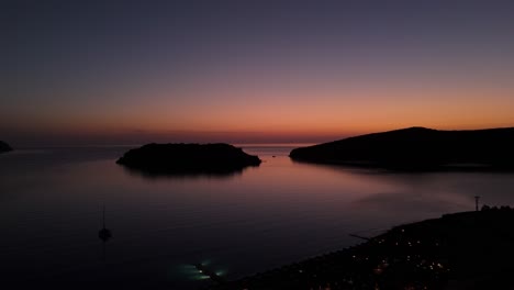 Aerial-ascend-above-Spinalogka-Greece-as-coastal-town-lights-flicker-with-blue-hour-sunset-glow-spreading-across-open-ocean-horizon